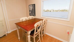 A dining room with a wooden table and white chairs, a window with blinds, and a framed picture on the wall. - Sunfish Sanctuary 2 Queen Deluxe at Fin N Feather (Nags Head)