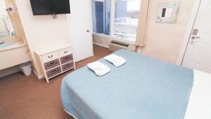 A hotel room with a bed, a TV mounted on the wall, a window with a view, and a small cabinet with drawers. - Sunfish Sanctuary 2 Queen Deluxe at Fin N Feather (Nags Head)