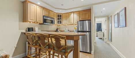 A modern kitchen with wooden cabinets, a stainless steel refrigerator, and a bar area with wicker chairs.