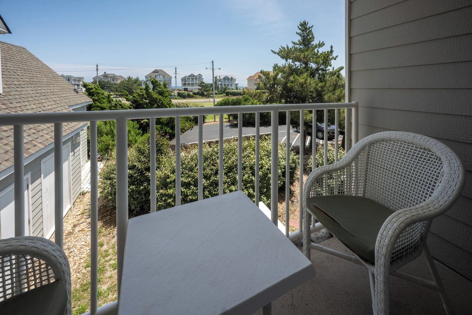 A balcony with a white table and two wicker chairs, overlooking a residential area with houses and a road.
