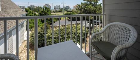 A balcony with a white table and two wicker chairs, overlooking a residential area with houses and a road.