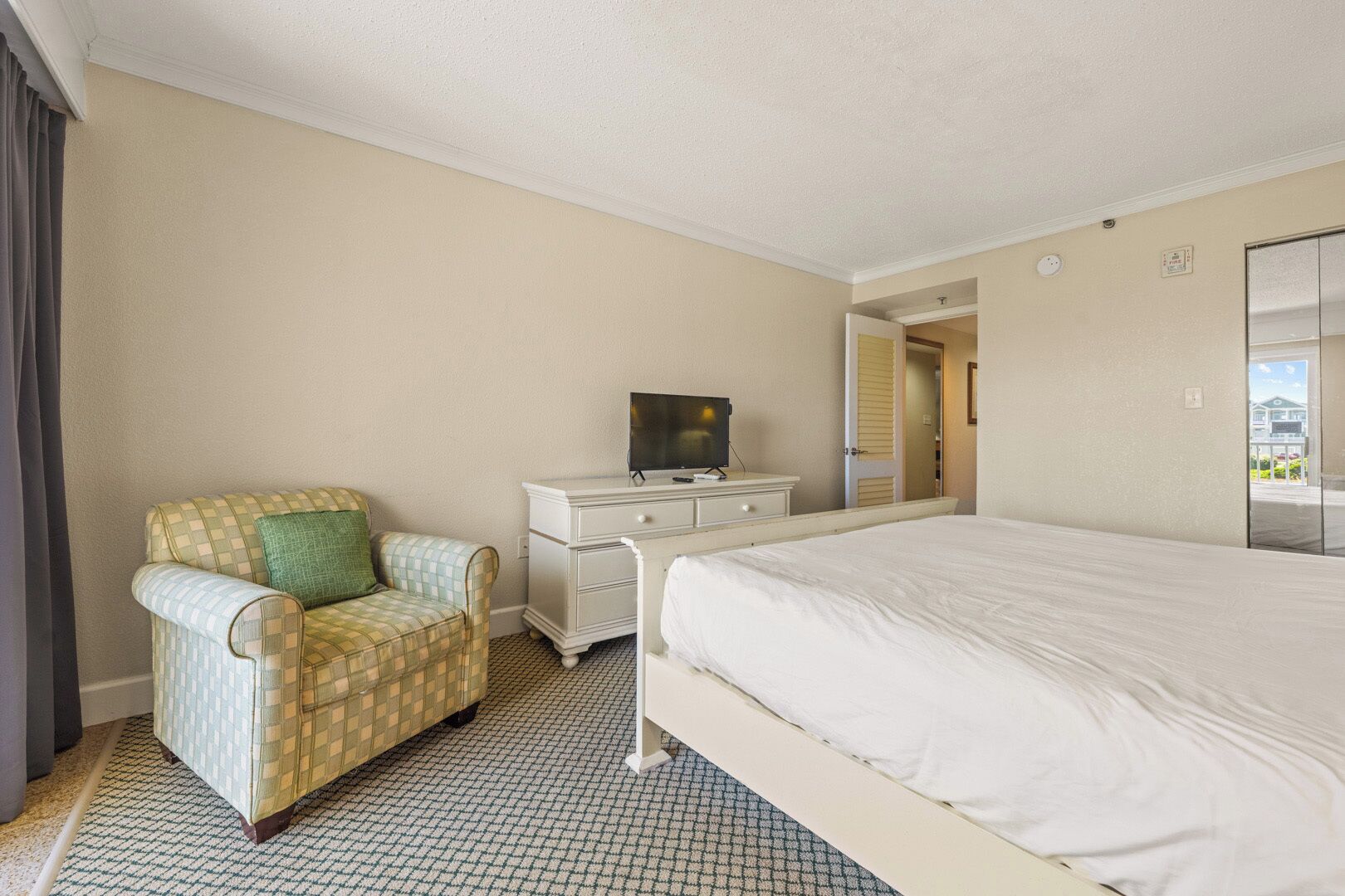 A hotel room with a checkered armchair, a white dresser with a television, a large bed, and a view through an open door.