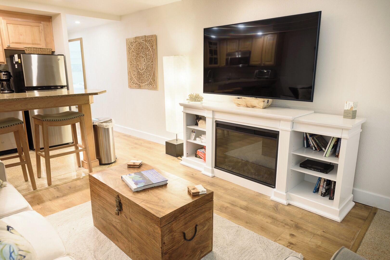 A modern living room with a wooden coffee table, a flat-screen TV mounted above a fireplace, and a kitchen area in the background.