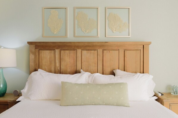 Un lit bien fait, avec du linge de lit blanc et un seul coussin décoratif, une tête de lit en bois et trois œuvres d’art encadrées accrochées au mur au-dessus.