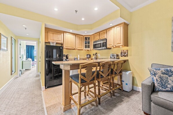 A compact kitchen with wooden cabinets, a black refrigerator, a microwave, and a small island with bar stools. - Get To The Point at the Villas at Hatteras Landing 1 Bedroom Near-Beach Condos with Pool Access! (Hatteras)