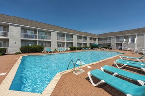 A swimming pool with lounge chairs, a brick patio, and a multi-story building with balconies.
