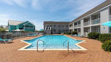 A swimming pool with a brick deck, surrounded by lounge chairs and a building with balconies.