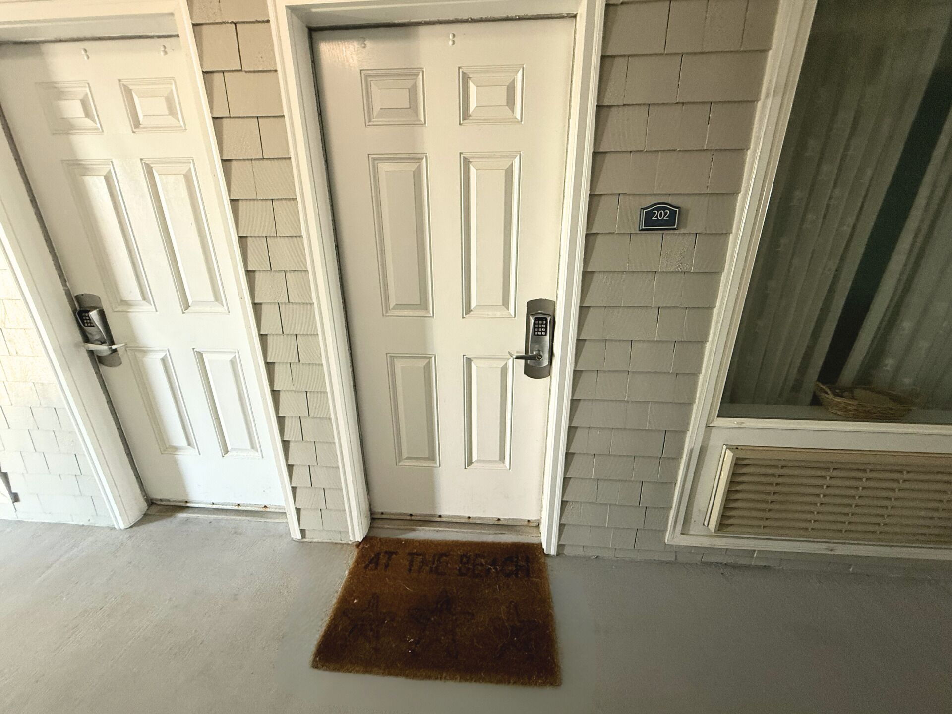 A set of white doors with a keypad lock, a doormat with the text "AT THE DOOR," and a house number sign.