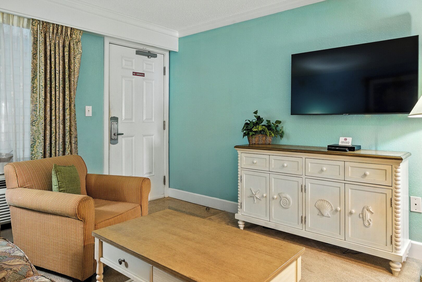 A living room with a sofa, a wooden coffee table, a white cabinet with decorative shells, and a television mounted on the wall.