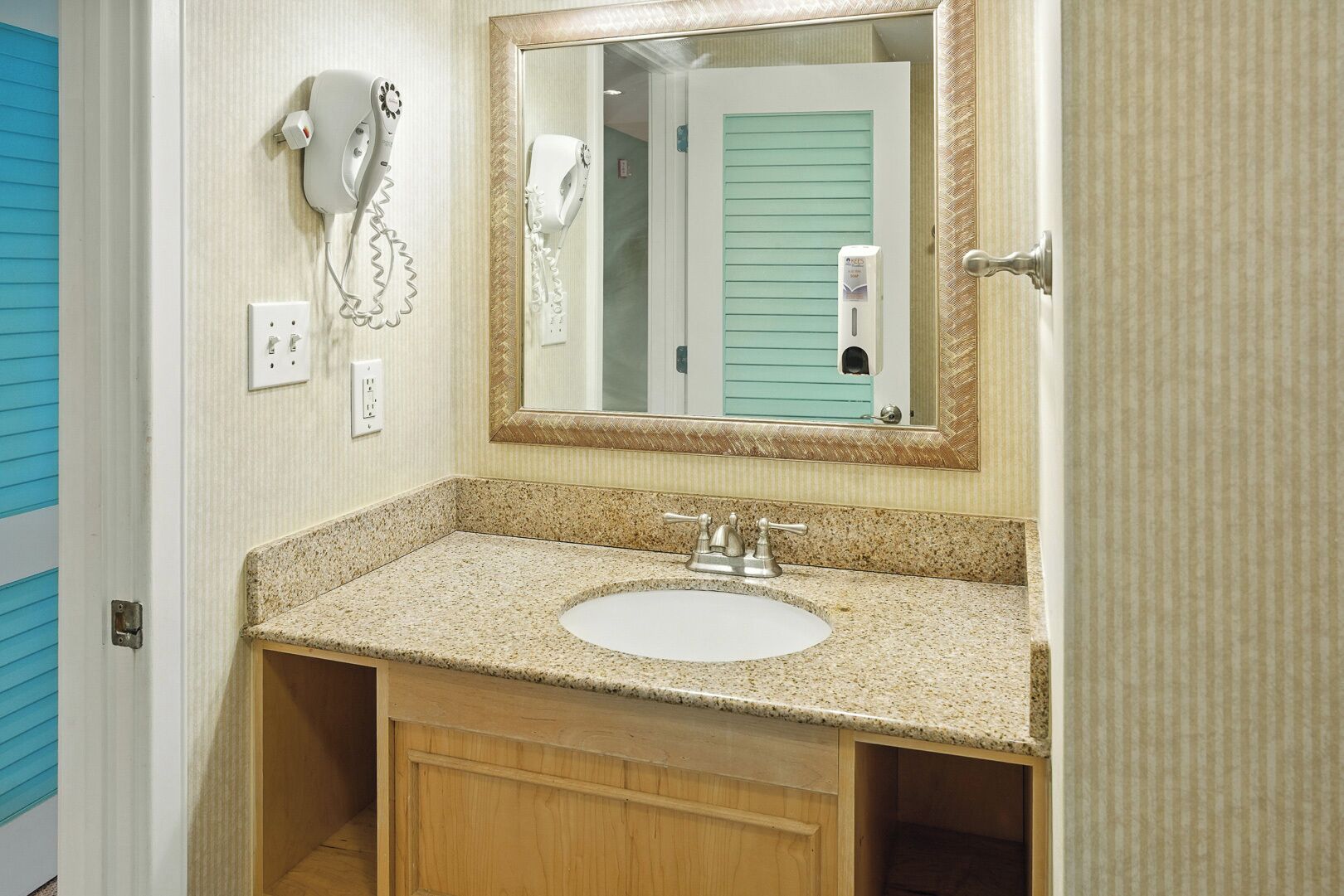 A bathroom with a granite countertop sink, a mirror with a gold frame, and two hair dryers mounted on the wall.
