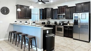 Una cocina moderna con armarios oscuros, electrodomésticos de acero inoxidable, una isla central con taburetes y un ventilador de techo.