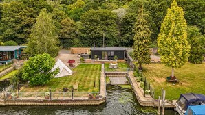 A lakeside property with a small dock, a white tent, a deck with outdoor furniture, and a building with a dark roof.