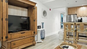 Un salon moderne avec un meuble de rangement en bois pour l’équipement audiovisuel, un grand téléviseur à écran plat, des fauteuils en osier et un coin cuisine doté de meubles en bois et d’appareils électroménagers en acier inoxydable.