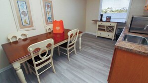 Un comedor con mesa y sillas de madera, una pequeña cocina con pileta y cafetera, y cuadros en la pared.