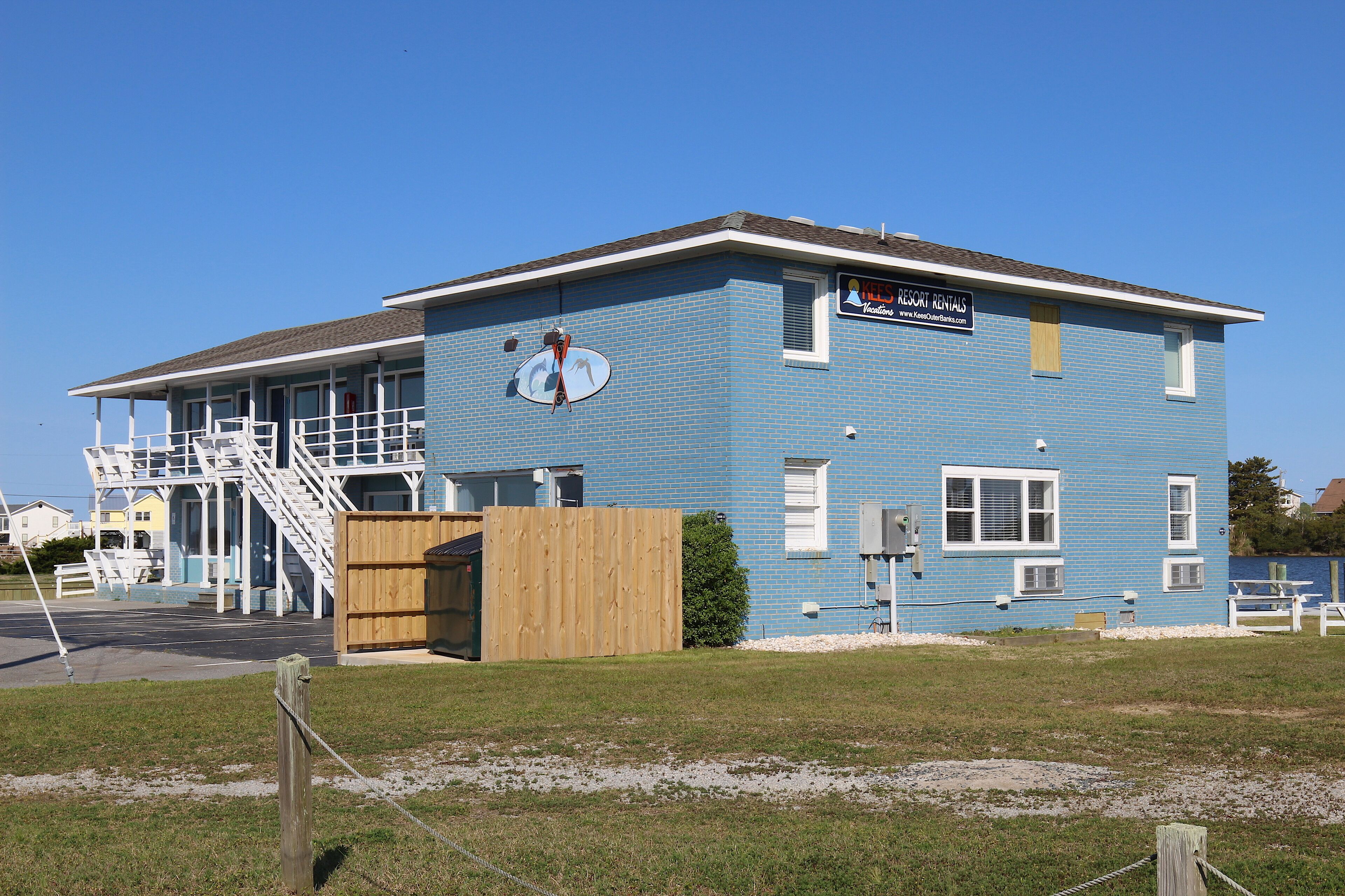Een gebouw met twee verdiepingen, een blauwe gevel, een houten terras en een bord waarop staat dat het een vakantiewoning is.