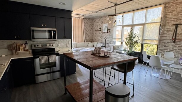 A modern kitchen with dark cabinets, a wooden island, stainless steel appliances, and a large window providing natural light.
