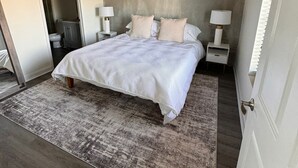 A bedroom with a large bed, two bedside tables with lamps, a concrete accent wall, and a wooden floor.