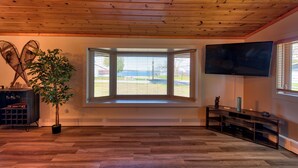 Quarto com janela grande, piso de madeira, planta em vaso, televisão e móvel de entretenimento de madeira.