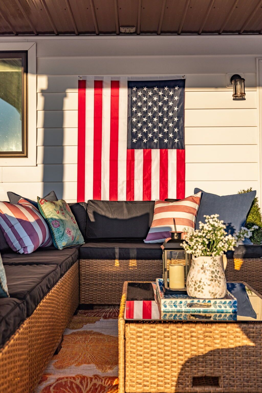 En hyggelig udendørs siddekrog med en sofa, puder, et flettet sofabord og et stort amerikansk flag.