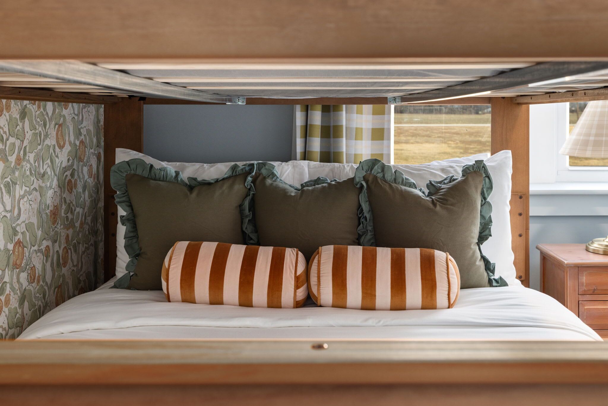 A bed with striped pillows and ruffled edges, a patterned wall, and a window with curtains.