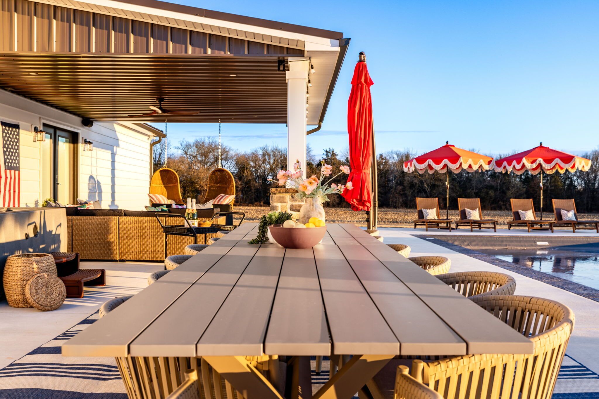 Een overdekt terras met eethoek, een zwembad, rieten meubels en rode parasols.
