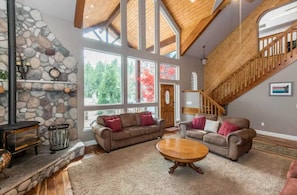 A spacious living room with a stone fireplace, two sofas, a coffee table, and a wooden staircase. - Handels Pride- Family friendly, Game Room & Views (Shaver Lake)