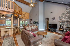 A spacious living area with a high ceiling, wooden beams, a stone fireplace, and a large TV.