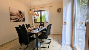 Ein Essbereich mit einem für vier Personen gedeckten Tisch, schwarzen Stühlen, hängenden Pendelleuchten, einem Gemälde an der Wand und einem Fenster mit Blick auf Grünflächen.