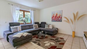 Ein modernes Wohnzimmer mit einem grauen Ecksofa, einem Glastisch und einem großen Fenster mit weißen Vorhängen.