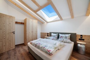 Un dormitorio con tragaluces, vigas de madera, una cama con ropa de cama floral, una mesita de noche y un cesto de basura.