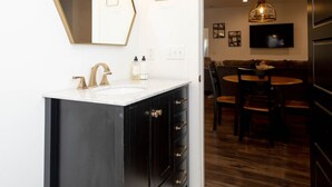 Ein modernes Badezimmer mit einem schwarzen Waschtisch, einem weißen Waschbecken und einem großen Spiegel. Ein Mülleimer und ein weißer Teppich liegen auf dem Boden.