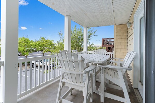 A covered outdoor patio with white Adirondack chairs and a table, overlooking a street with parked cars and trees. - Spacious 3-Bedroom Ocean Block Townhome – Steps from the Sand! (Ocean City)