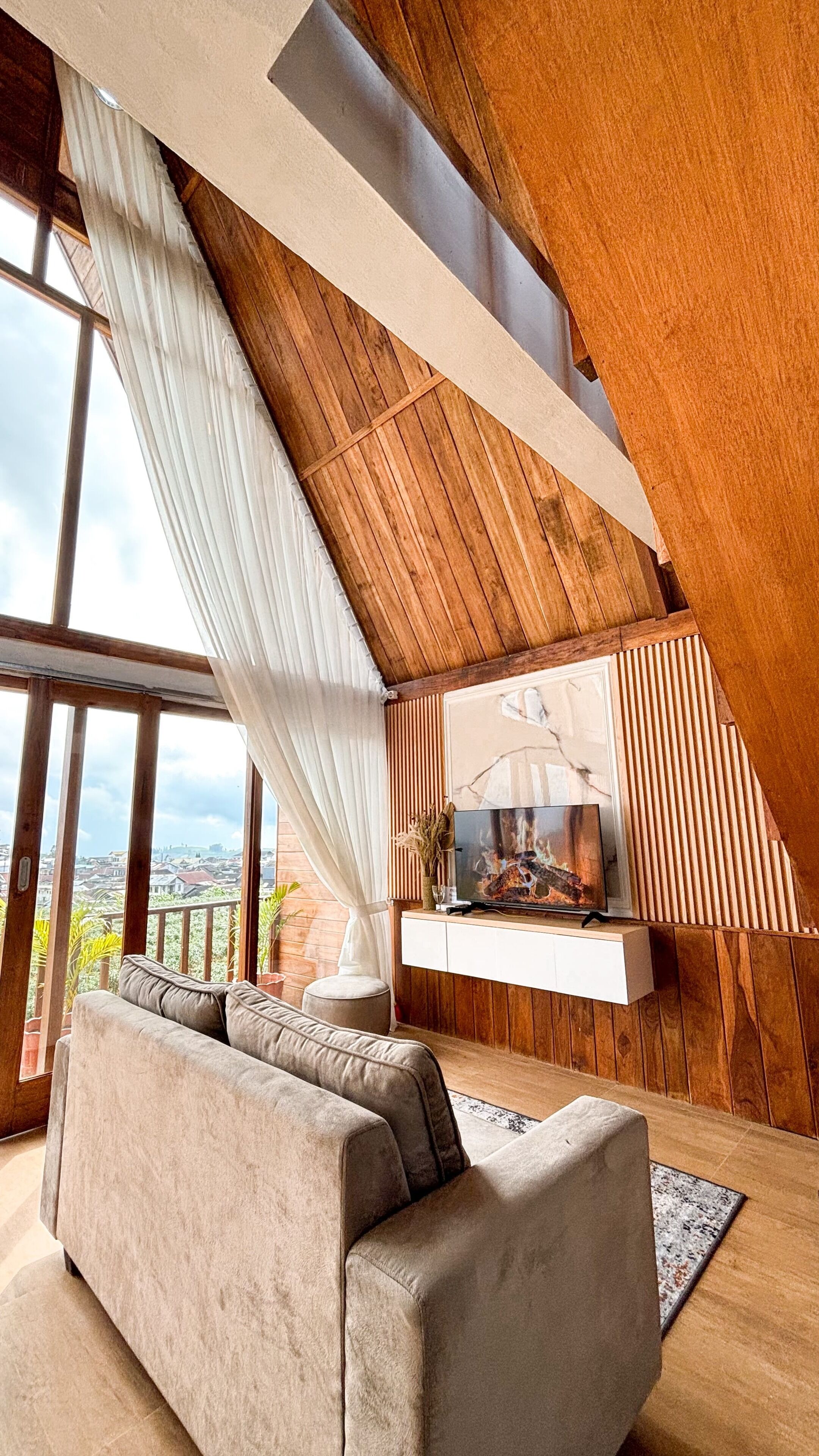 Ein modernes Wohnzimmer mit hohen Decken, Holzbalken, einem großen Fenster mit Vorhängen, einer Couch und einem flachbildfernseher, der an einer Holzwand angebracht ist.