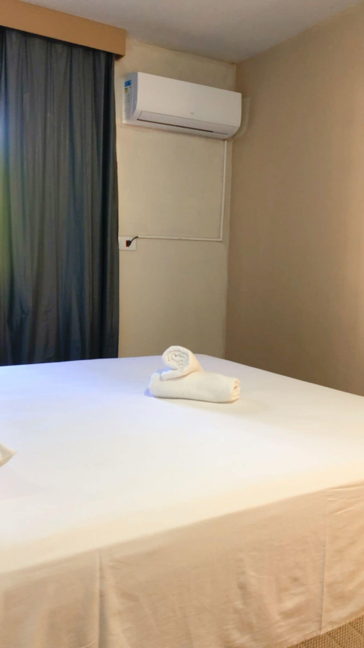 A hotel room with a bed, a rolled towel, an air conditioning unit, and a curtain.