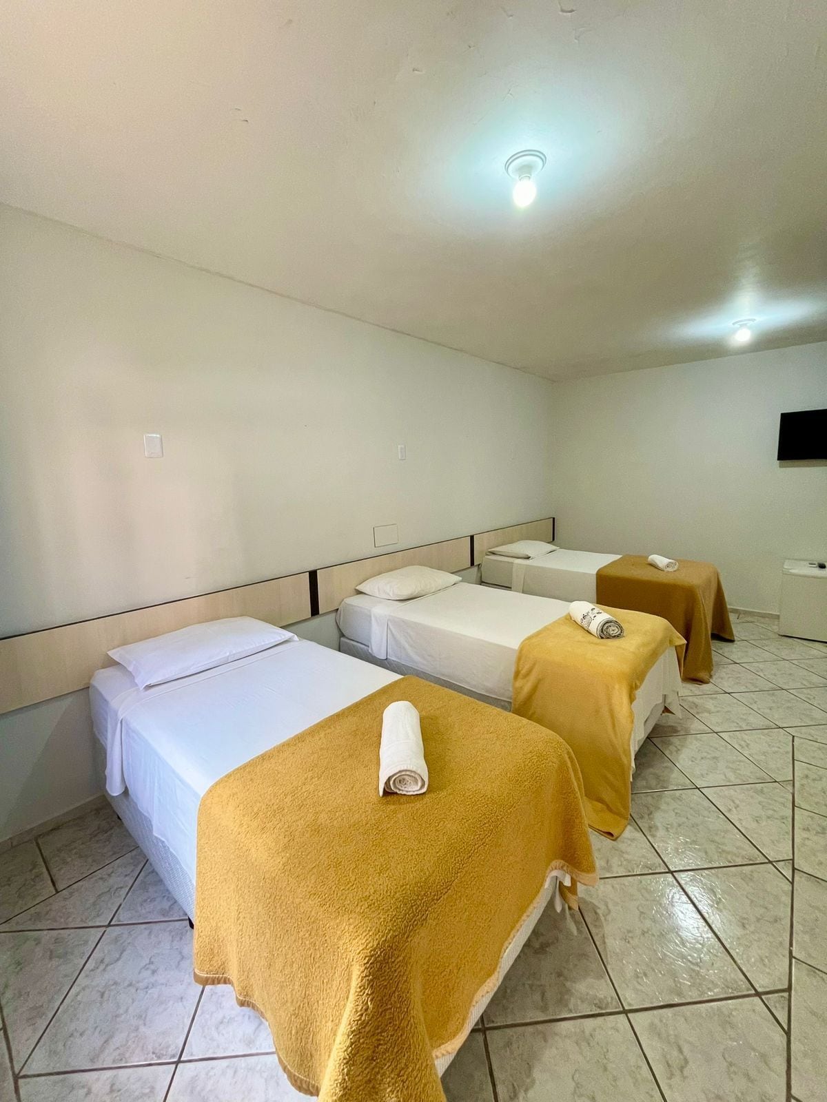 A hotel room with three beds, each with a white bedspread and a rolled towel on the headboard.