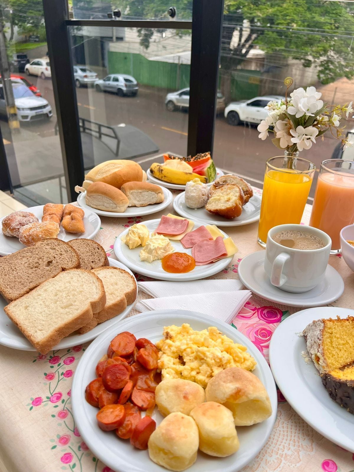 A breakfast table with various food items and drinks, a window view of a street, and a vase with flowers.