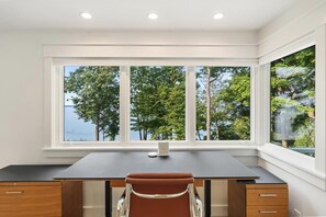 A room with a desk, chair, and large windows offering a view of trees and a body of water.