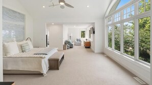 Une chambre avec un grand lit, un ventilateur de plafond et une vue sur les arbres à travers de grandes fenêtres.