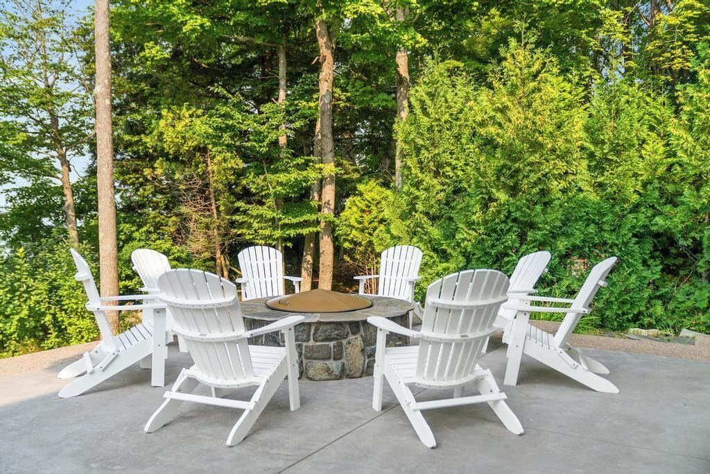 Een rond zitgedeelte buiten, met witte Adirondack-stoelen rond een vuurkorf van steen.