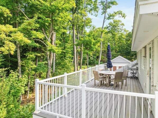 A wooden deck with a white railing, surrounded by trees, and a table with chairs and an umbrella.
