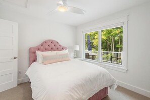 A bedroom with a bed, a ceiling fan, a window with a view of trees, and a door.