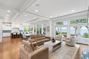 A spacious living room with a large window overlooking a body of water, a kitchen with white cabinetry, and a dining area with a table and chairs. - Blue Beech Estate- Beautiful Waterfront Estate! (Harbor Springs)