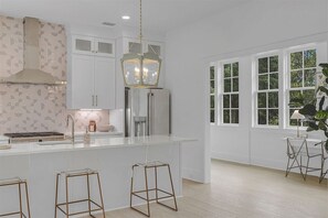 A modern kitchen with white cabinetry, a central island with stools, a stainless steel refrigerator, and a large window with a view of greenery.