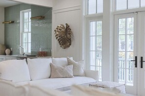 A white sofa with decorative pillows, a large leaf wall art, and a window with a view.