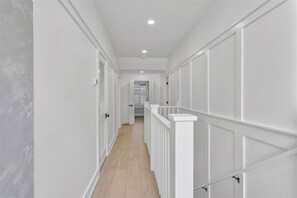 Een smalle gang met witte muren en houten vloeren, die leidt naar een kamer met een raam en een deur.