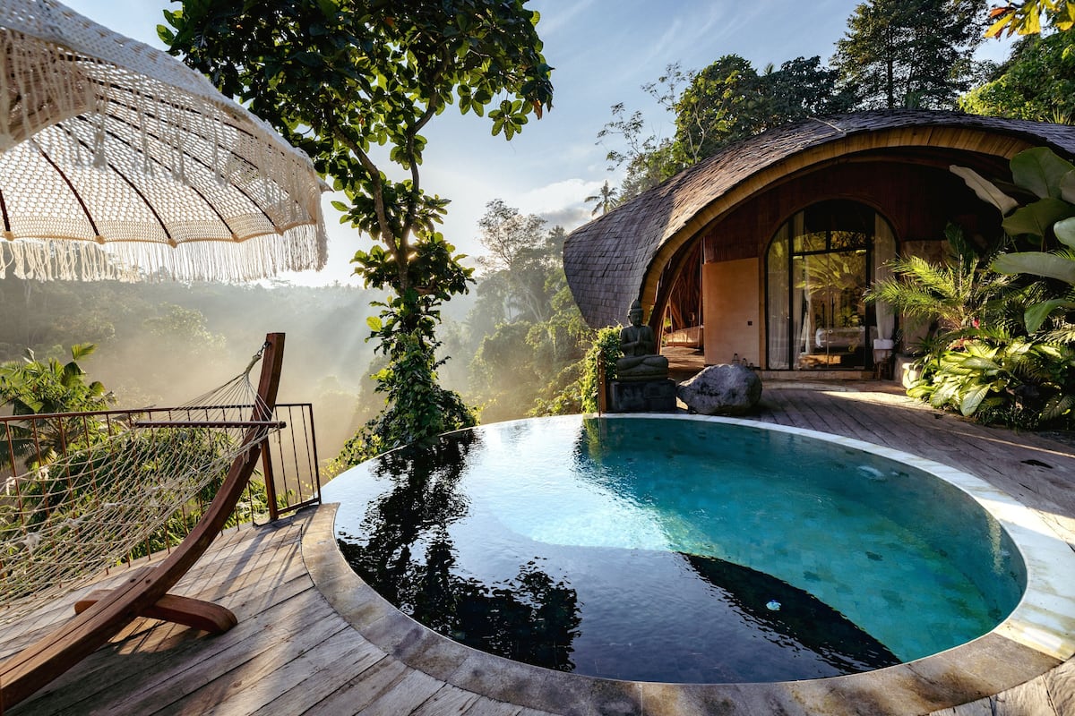 Une terrasse en bois avec une piscine, entourée d’une végétation luxuriante et d’une structure à toit de chaume.