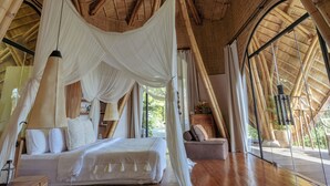 A spacious room with a high, curved bamboo ceiling, a large bed with white bedding, and a view through a large window. - Belalu Bali Boutique & Spa Experience (Ubud)