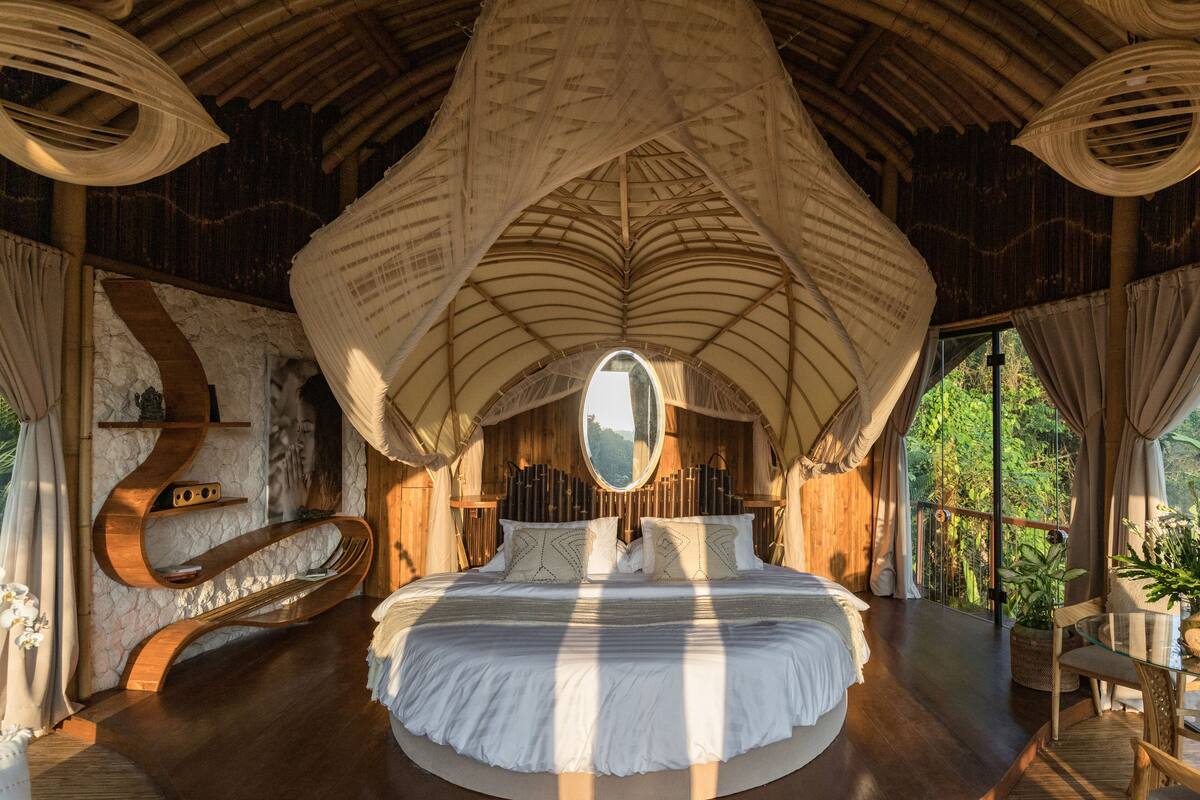 Une chambre spacieuse avec un grand lit, un banc en bois aux lignes courbes, un miroir rond et une vue sur la verdure grâce à de grandes fenêtres.