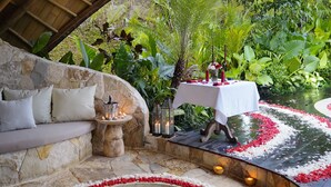 A stone-walled outdoor seating area with a round hot tub surrounded by red and white flower petals, a table with candles, and lush greenery in the background. - Belalu Bali Boutique & Spa Experience (Ubud)