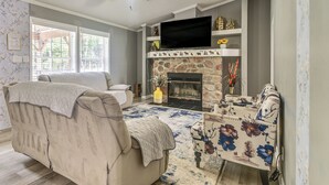 A living room with a beige sofa, a floral armchair, a fireplace, a television, and a ceiling fan.